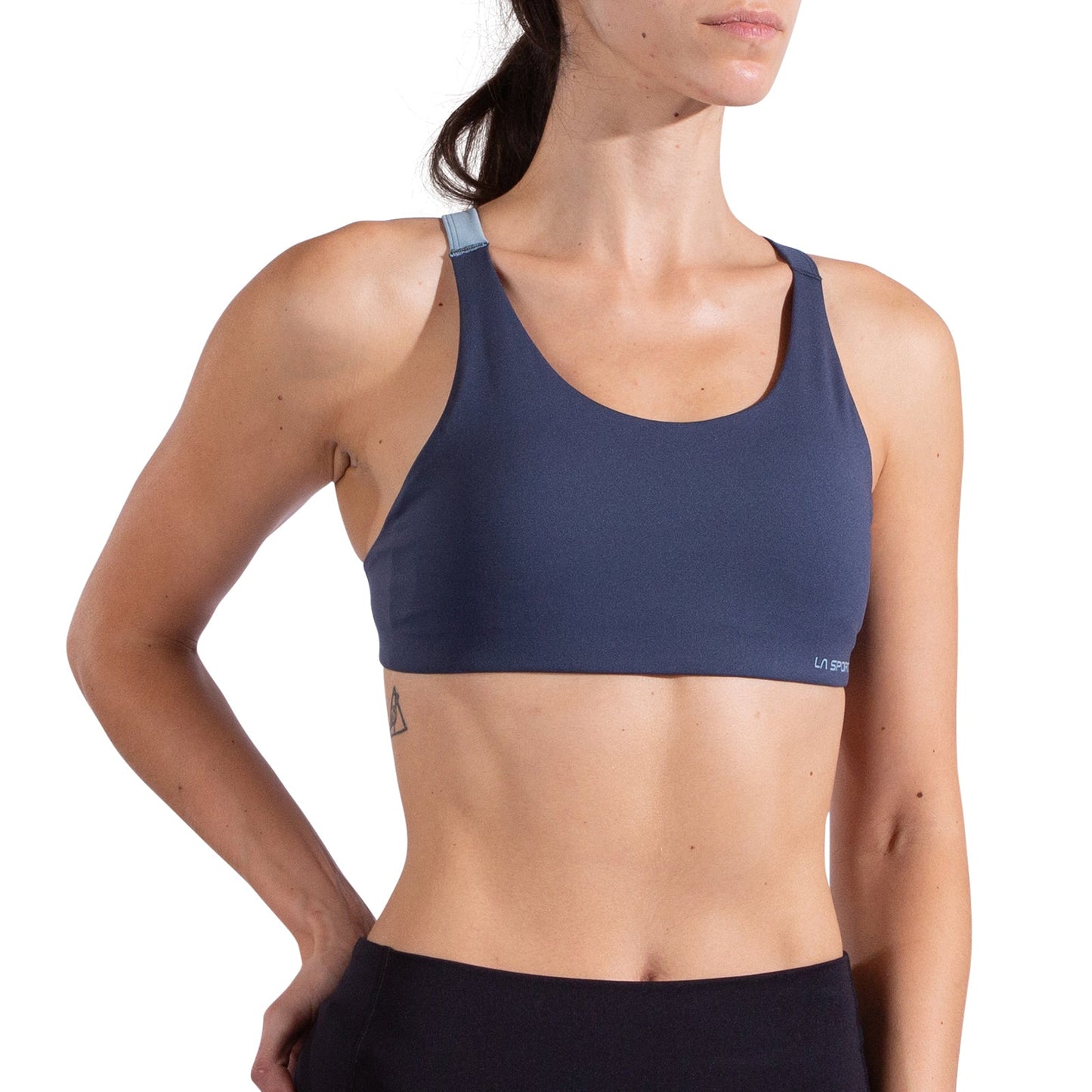 A woman models the La Sportiva Cosmosis Bra Women's in dark blue with light blue adjustable straps, displaying a small left-side tattoo and standing with one hand on her hip; only her upper body is visible.