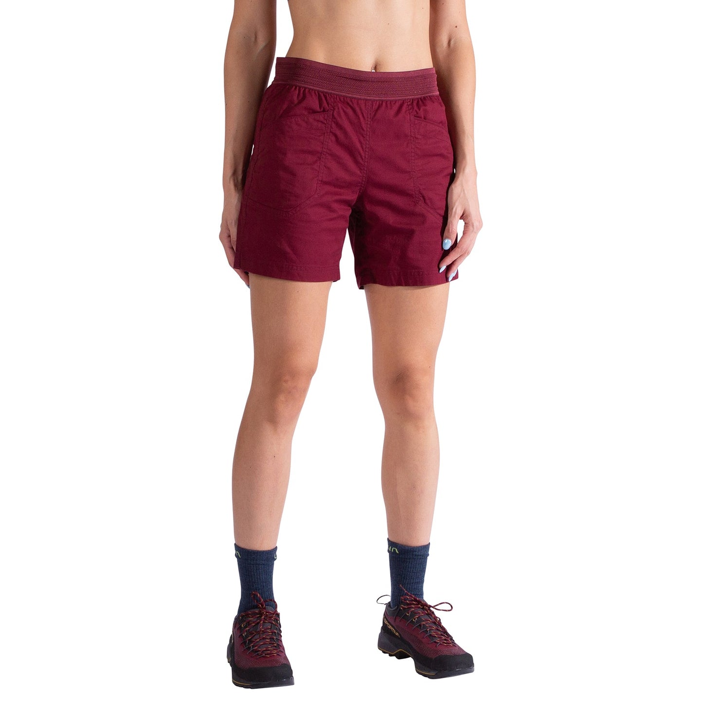 A woman wears La Sportiva Roots Shorts Women's in maroon organic cotton with durable pockets, paired with navy socks and hiking shoes, standing arms relaxed and face out of frame against a plain white background.