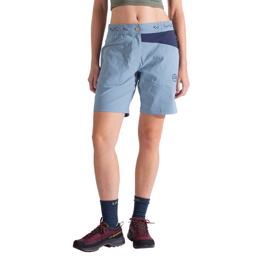 A person wearing La Sportiva Mantra Shorts Women's in light blue recycled fabric with a dark blue waistband, button closure, and chalk bag loop stands against a white background; navy socks and hiking shoes complete the look. Upper body is partially out of frame.