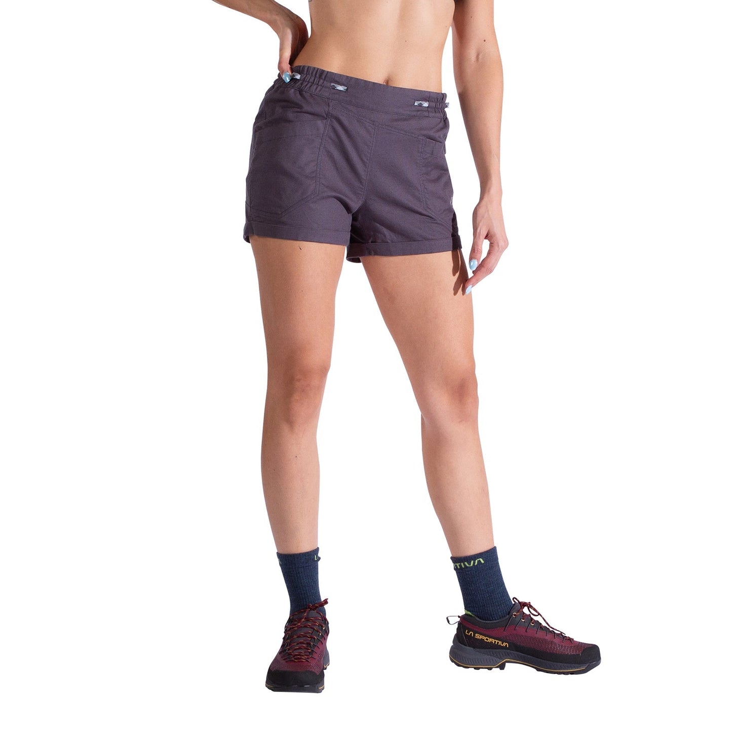 A person stands against a white background wearing La Sportiva Bolt Shorts Women's in dark gray with an adjustable waistband, black socks, and burgundy trail shoes. Their torso is partially visible, one arm on their hip, the other relaxed.