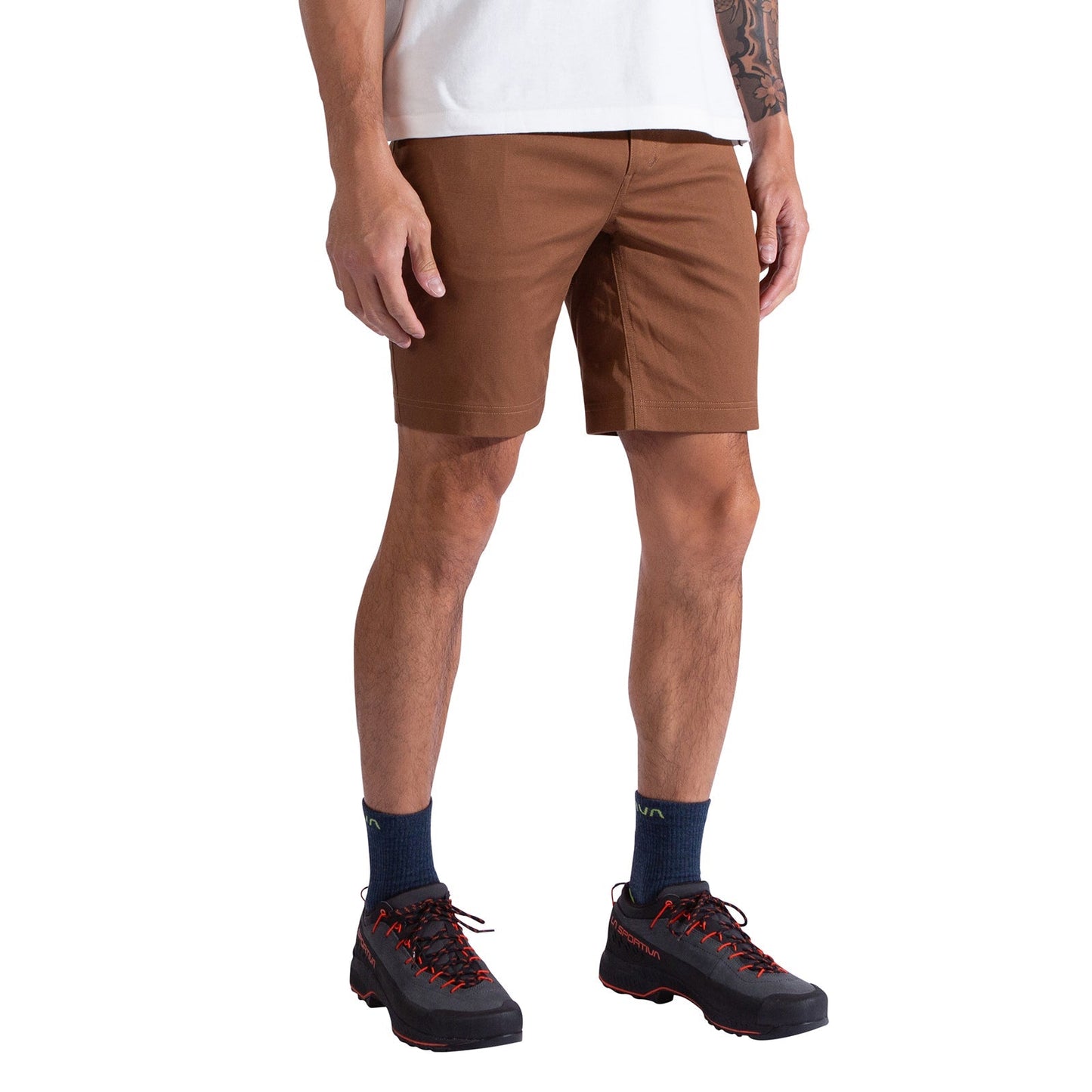 A person stands against a white background wearing a white T-shirt, navy socks, black hiking shoes with red laces, and La Sportiva Mainliner Shorts Men's made from durable organic cotton in brown.