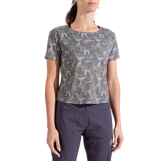A woman stands against a white background wearing the La Sportiva Dimension T-Shirt Women's, a gray geometric-patterned short-sleeve shirt made from recycled polyester with a subtle chest logo, paired with dark pants for an eco-friendly look.