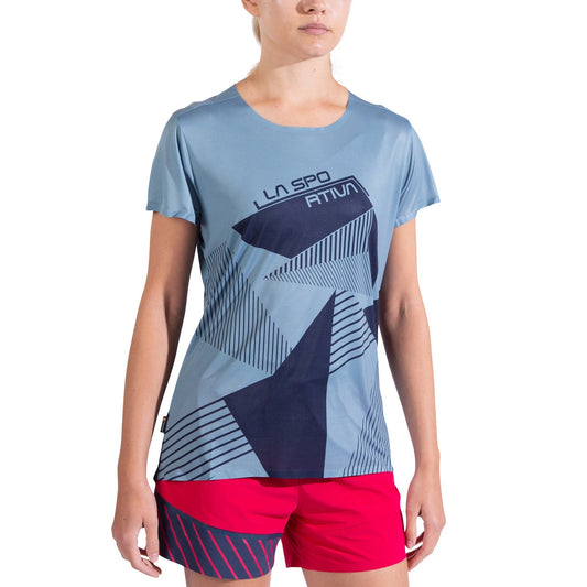 A woman models the La Sportiva Comp T-Shirt Women's, a lightweight light blue top with geometric navy patterns, paired with red shorts featuring diagonal navy stripes. Her face is partially out of frame against a white background.