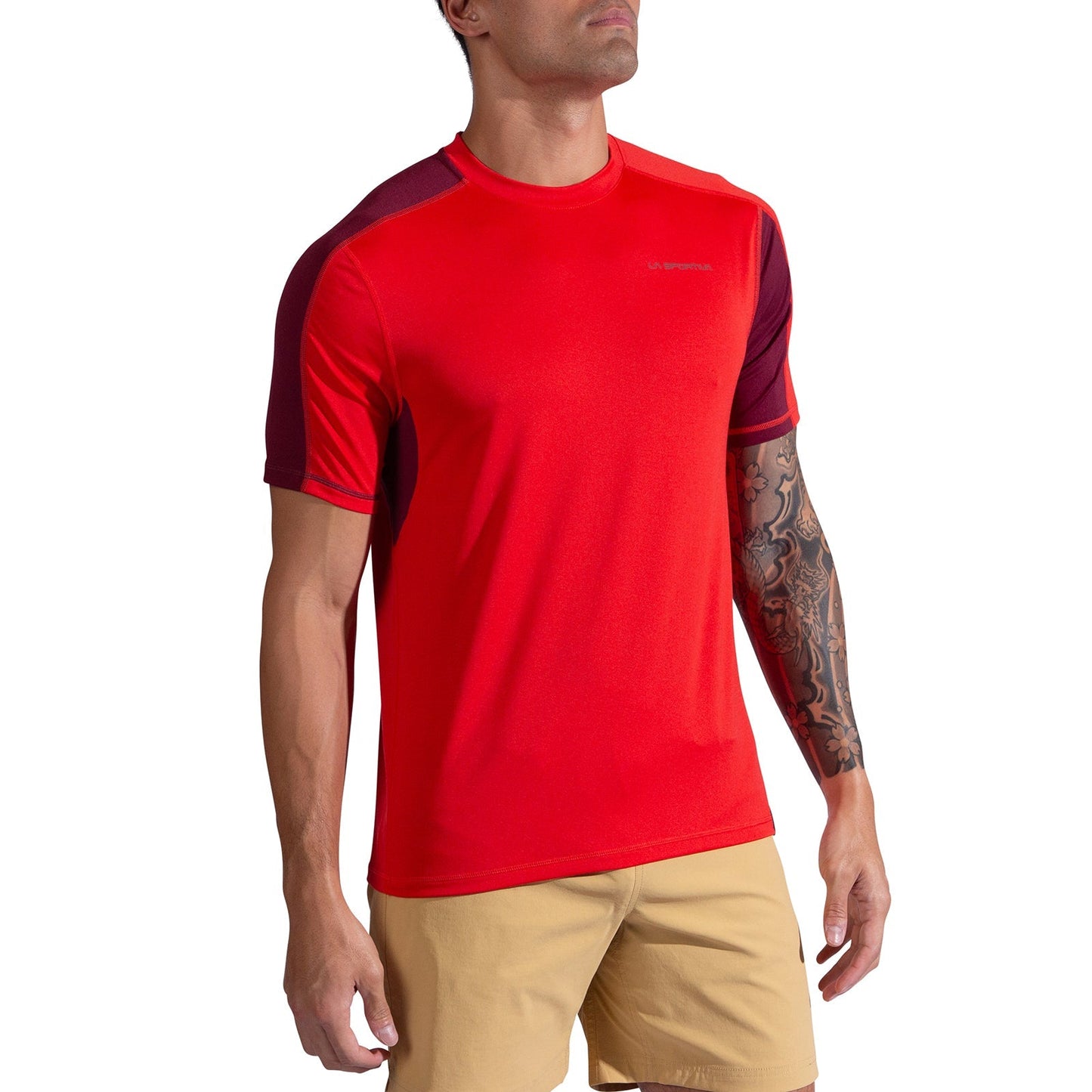 A man wearing the La Sportiva Just Right T-Shirt Men's in bright red with maroon sleeve detail and tan shorts, showing a tattoo on his right forearm. He stands slightly angled, head tilted upward.