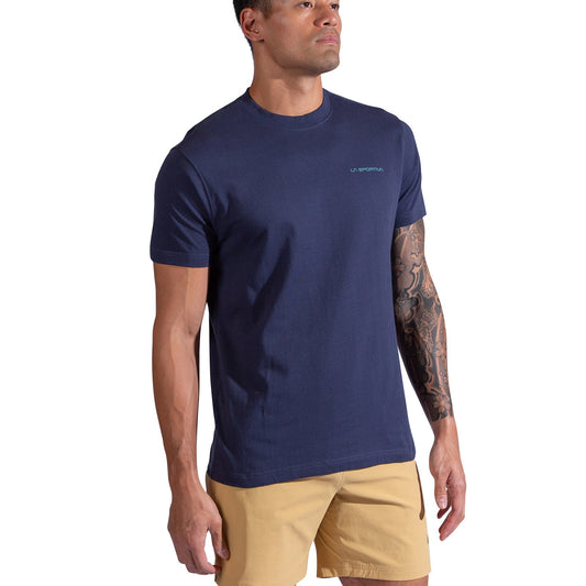 A man in a navy blue La Sportiva Psychotomic T-Shirt and tan shorts stands against a white background; the T-shirt features a small La Sportiva logo on the chest, and he has a tattoo on his left arm.