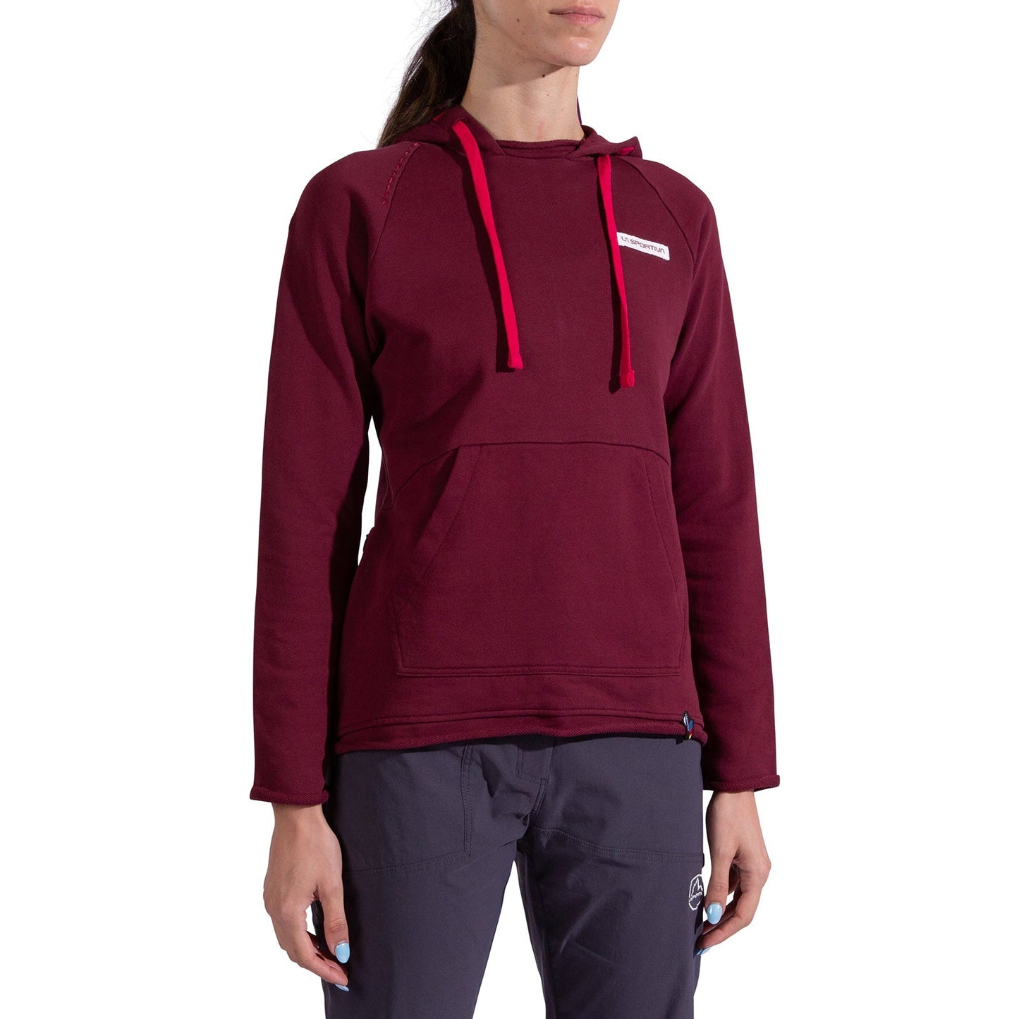 A woman models the La Sportiva Telendos Hoody Women's in burgundy with bright red drawstrings and a front pocket, paired with dark gray pants. The image captures her from shoulders down, head slightly out of frame.