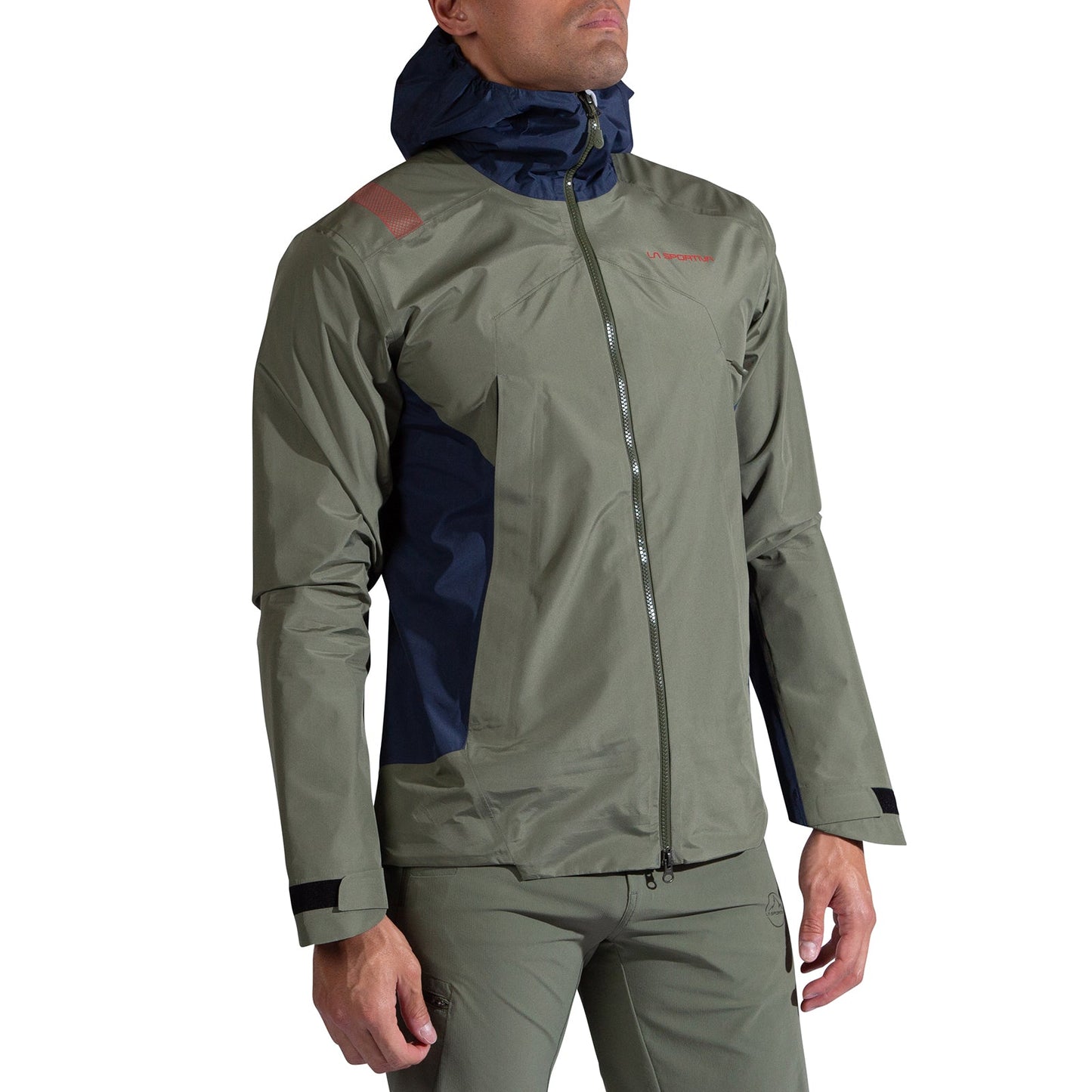 A man wears the La Sportiva Discover Shell Jacket Men's—olive-green with navy hood and side panels, front zipper, Velcro cuffs, and subtle chest branding—standing with arms relaxed.