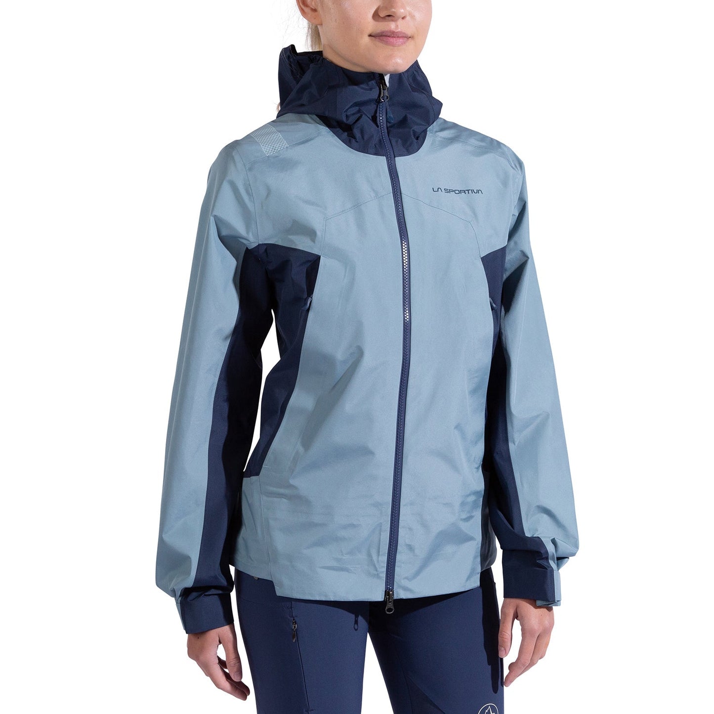 A woman models the La Sportiva Discover Shell Jacket in light blue and navy, zipped up with adjustable cuffs, paired with matching navy pants, against a white background.