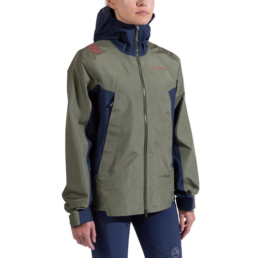 A woman wearing the La Sportiva Discover Shell Jacket in olive green and navy blue, featuring a front zipper and adjustable cuffs, stands against a plain white background.