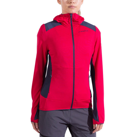 A woman wears the La Sportiva Aequilibrium Thermal Hoody in bright pink with dark gray accents and Polartec Power Grid fabric, featuring a front zipper and cuff thumb holes, paired with gray shorts. The background is plain white.