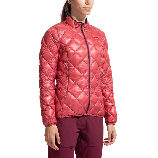 A woman models the La Sportiva Lumina 100 Down Jacket Women's—a shiny, quilted red lightweight down jacket with a front zipper—paired with maroon pants against a plain white background.