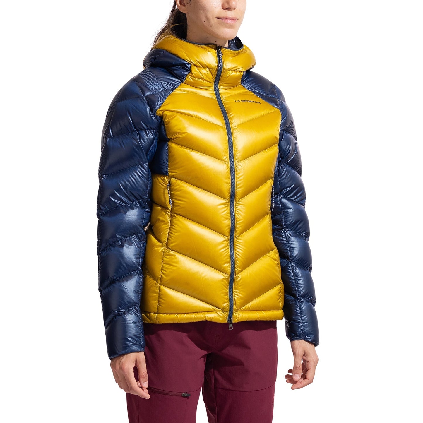 A person stands against a white background, wearing the La Sportiva Lumina 300 Down Jacket Women's with a shiny finish, yellow front, blue sleeves and shoulders, hood, and maroon pants.