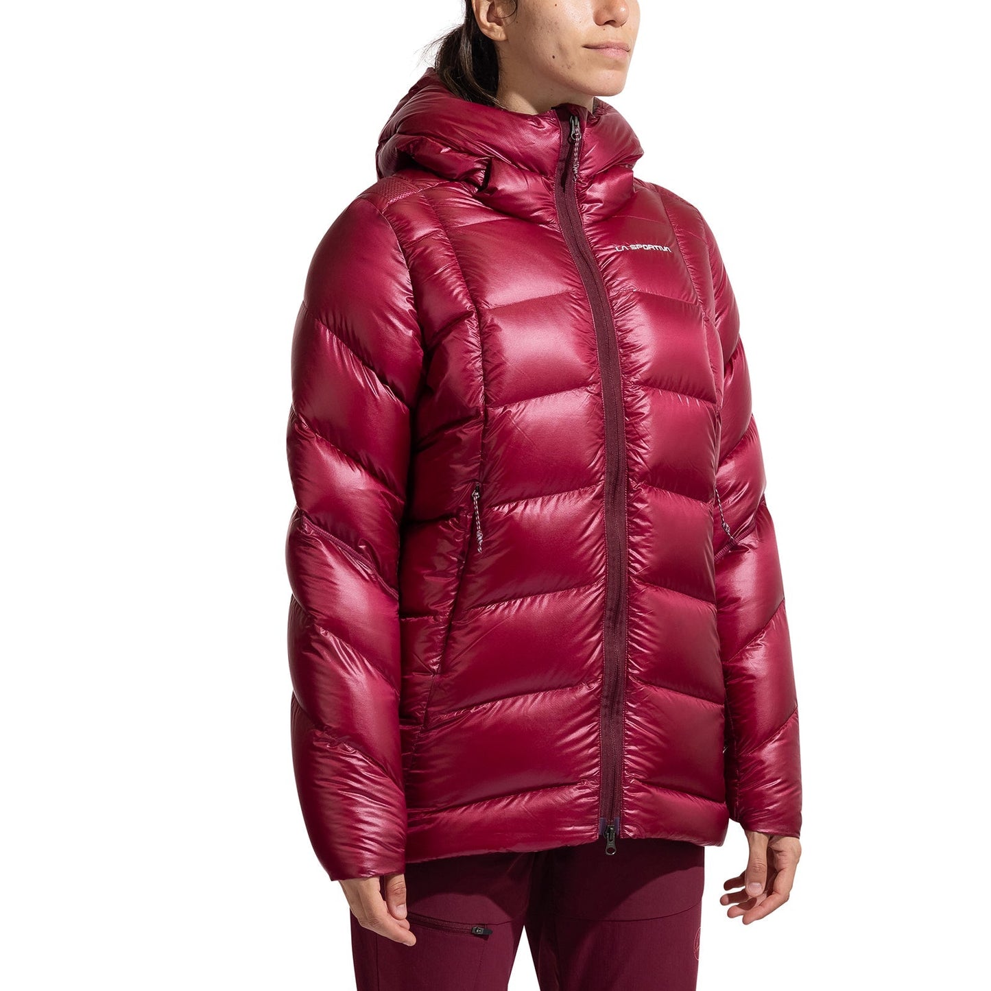 A woman models the La Sportiva Supercouloir Down Parka against a white background. The shiny maroon jacket features horizontal quilting, hood, front zipper, and is packable; her face is partially out of frame.