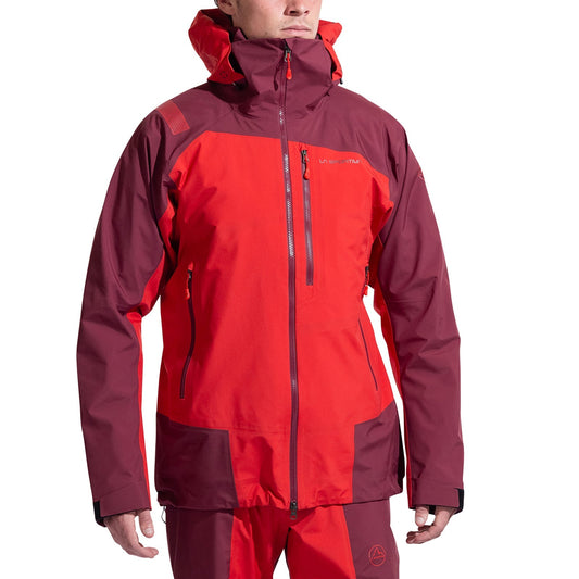 A person wears the La Sportiva Alpine Guide GTX Performance Jacket Men's in red and maroon, with matching pants. The jacket features multiple zippered pockets and adjustable cuffs, ideal for alpine and winter activities. Face is partially visible.