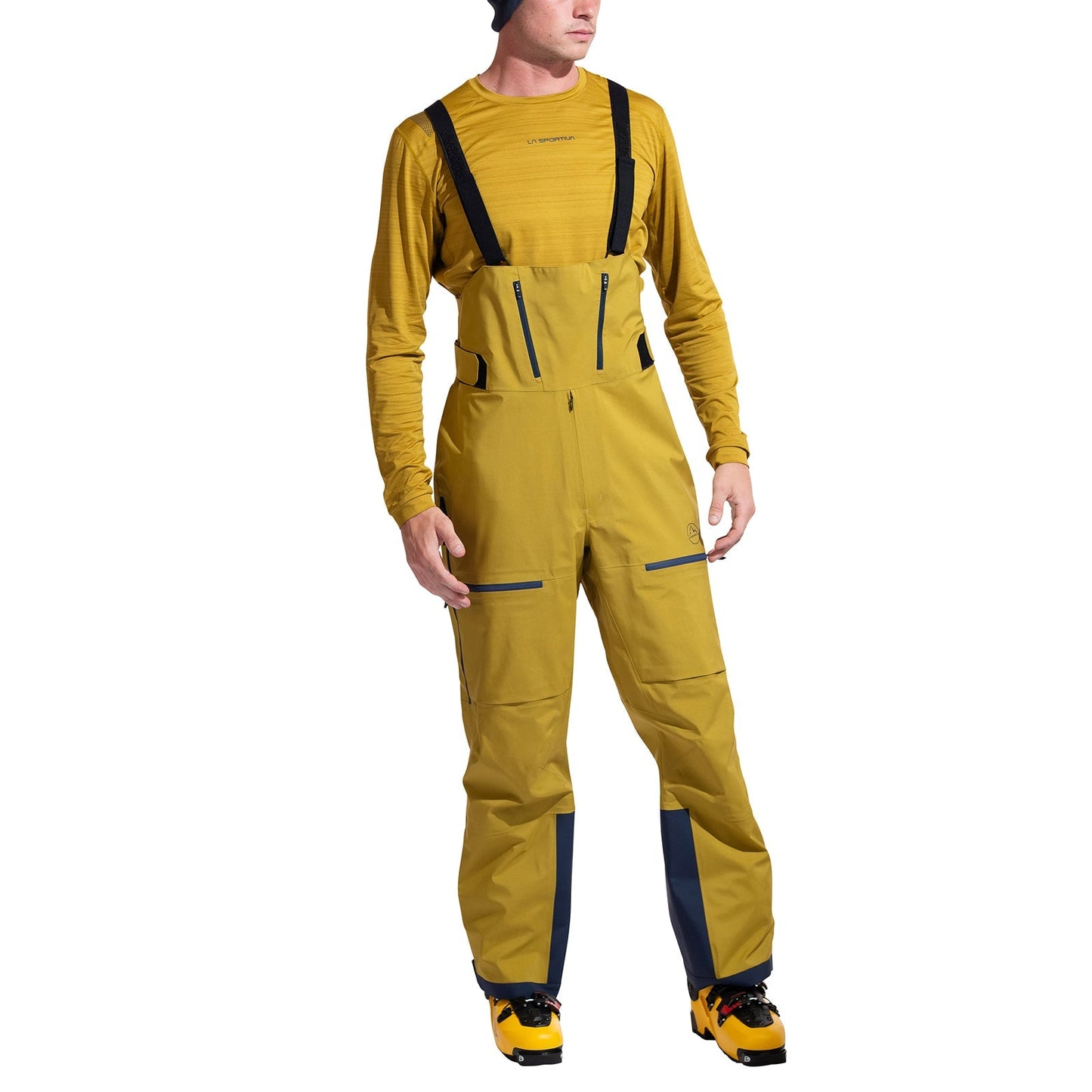 A person stands against a white background wearing a mustard yellow long-sleeve shirt, La Sportiva Sender GTX Performance Bib Pants Men's with black straps, and yellow and black ski boots.