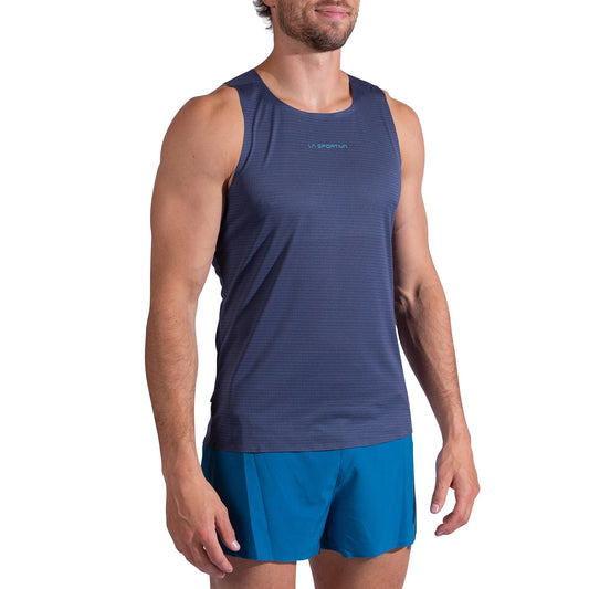 A man wears the La Sportiva Pure Tank Men's, a sleeveless navy trail running top with odor control, paired with bright blue shorts, standing with arms relaxed at his sides against a white background.