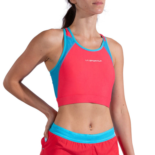 A woman wears the La Sportiva Stash Bra Women's, a medium-support, sleeveless crop top in red and blue with matching red shorts—perfect for trail runs. One hand rests on her hip and her face is partially out of frame.