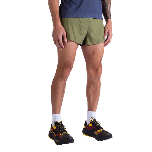 A person wearing La Sportiva Pure 3 Inch Shorts Men's in olive green stands against a white background. Only the lower half of the body is visible, paired with a navy blue top, white socks, and black and yellow running shoes.