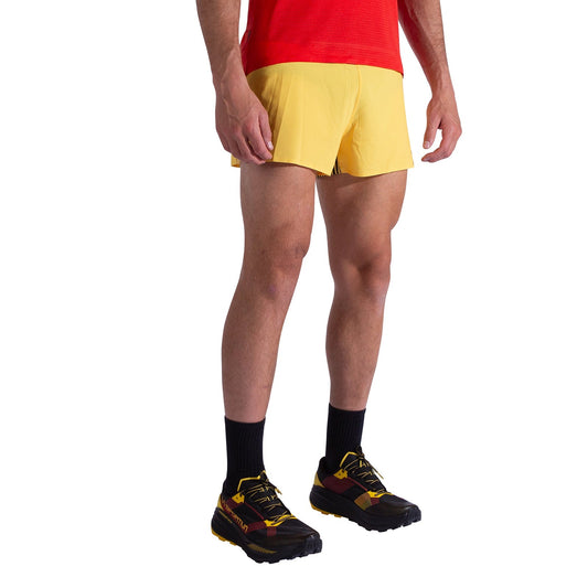 A man stands against a white background wearing La Sportiva Flow 3 Inch Shorts Men's in yellow, a red shirt, black socks, and black/yellow running shoes. His face is not visible.