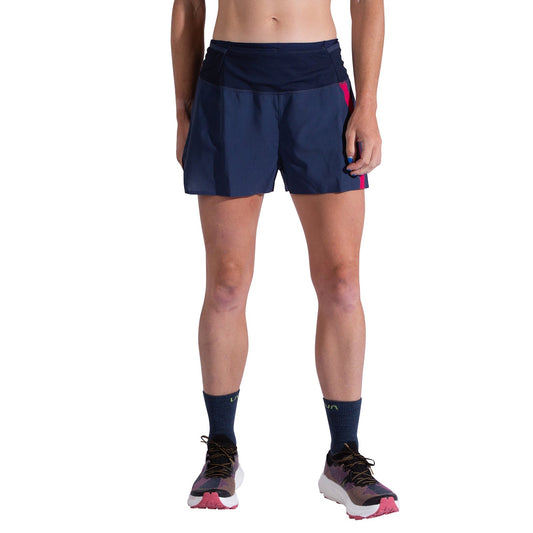A woman wears La Sportiva Flow 3 Inch Shorts in navy blue with a dark waistband and pink side accent, paired with tall navy socks and white running shoes with pink soles, standing against a white background.