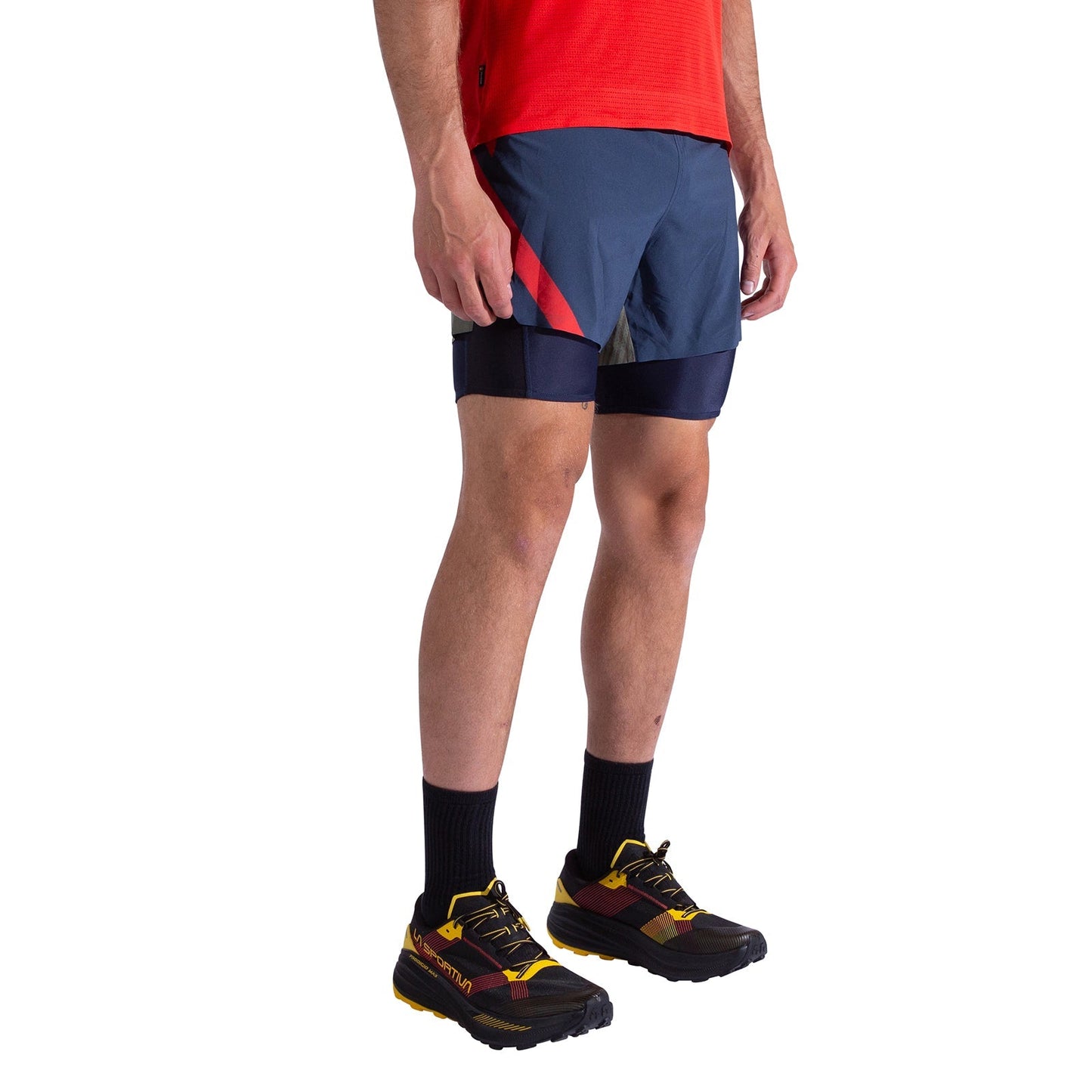 A person wearing a red shirt, La Sportiva Flow 5 Inch Shorts Men's in blue with red and black accents, black crew socks, and black trail running shoes with yellow and red details stands against a white background.