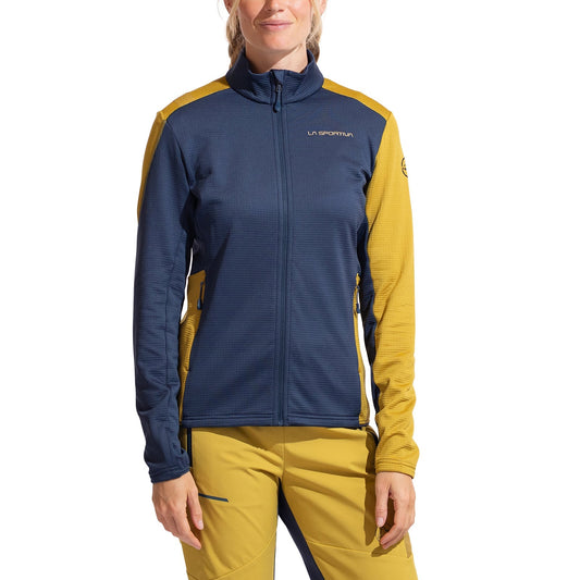 A woman wears the La Sportiva Chill Thermal Jacket Women's, featuring a dark blue front and sleeves with mustard yellow shoulders, arms, and sides. She pairs it with matching mustard yellow pants accented in blue.