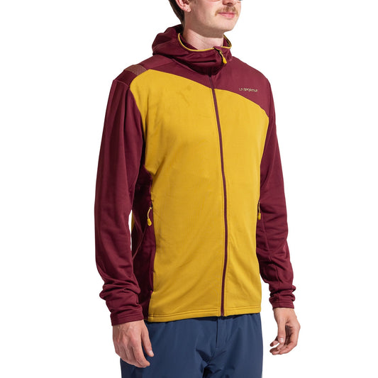 A man wears the La Sportiva Cosmic Thermal Hoody Men's in yellow and maroon with a front zipper and chest branding. Paired with blue pants, it's a stylish, functional choice for outdoor activities.