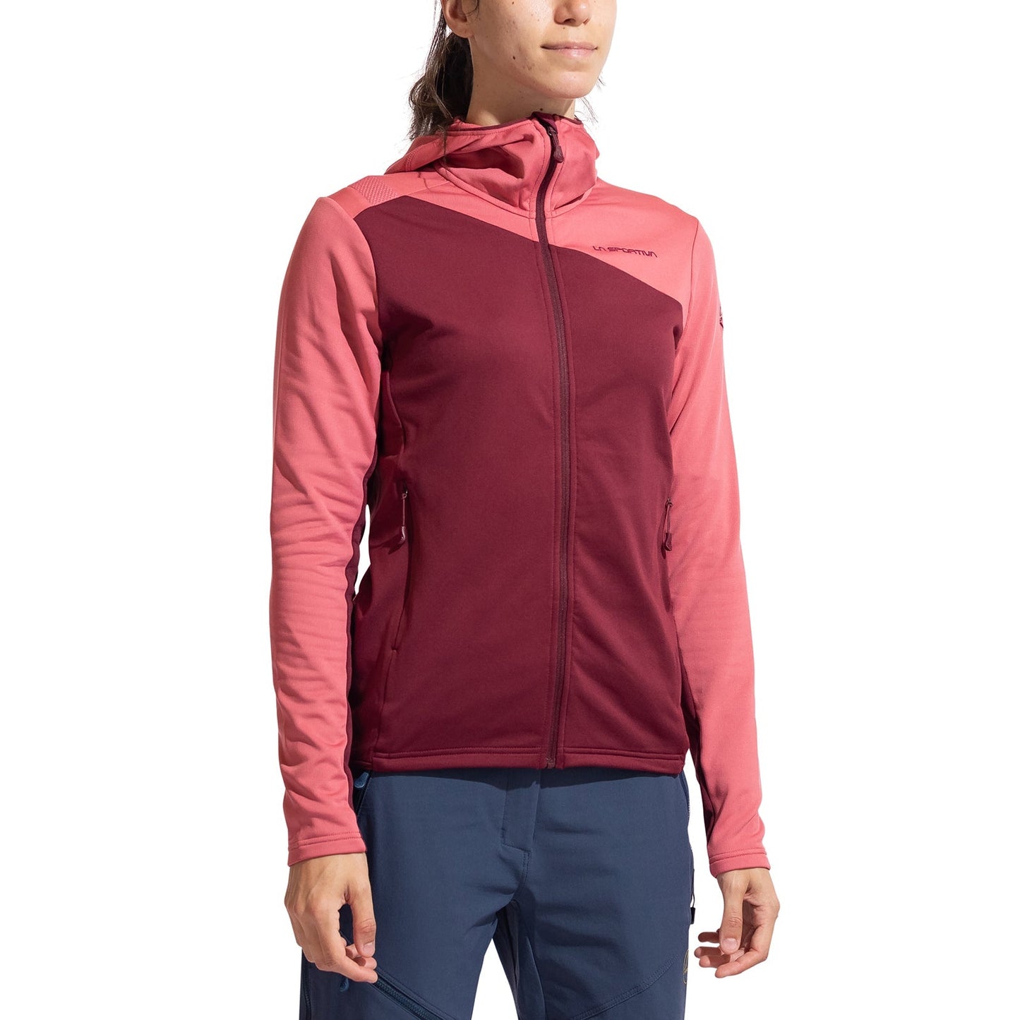 A woman wears the La Sportiva Cosmic Thermal Hoody Women's in two-tone pink and burgundy, made from Tecnostretch Pontetorto fabric, paired with blue pants. She stands against a white background, smiling softly and ready for outdoor adventures.