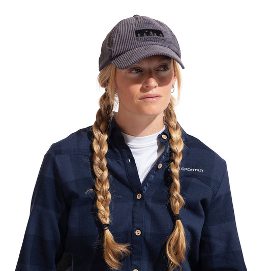 A woman with blonde braided hair wears the La Sportiva Setter Cap, a gray cotton corduroy climbing hat with an adjustable back buckle, paired with a blue plaid button-up shirt over a white top, looking slightly to the side against a white background.