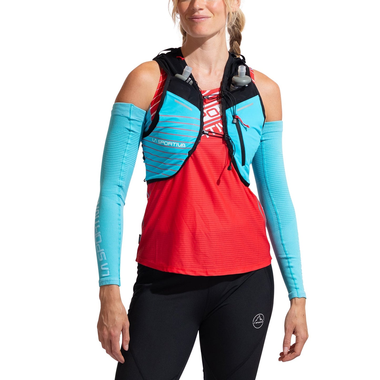 A woman ready for mountain running wears a red athletic top, La Sportiva Trace Arm Warmer in blue, black leggings, and a turquoise hydration vest with water bottles, standing against a white background.