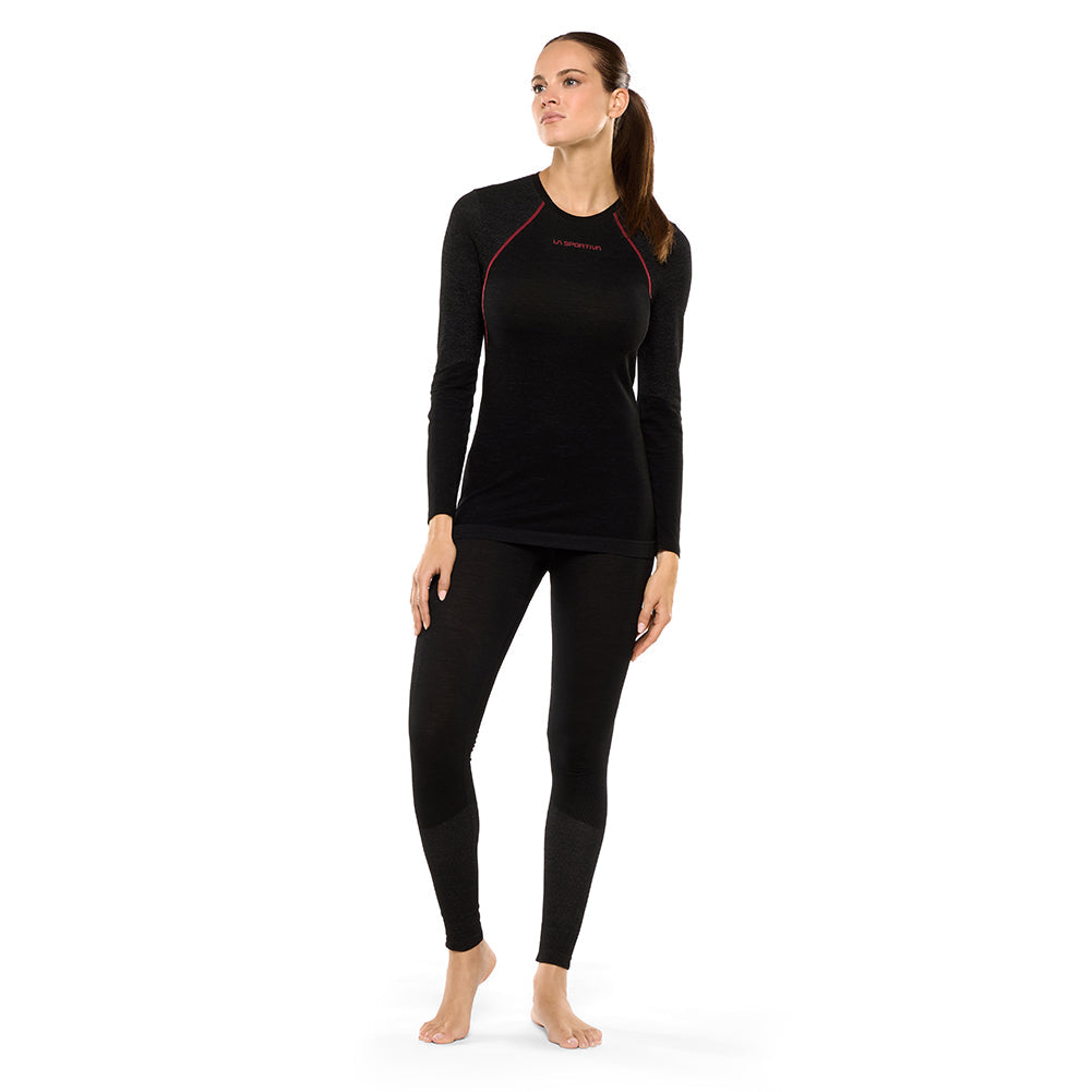 A woman stands barefoot against a white background, wearing the La Sportiva Wool40 Aero Long Sleeve Women's top and matching leggings—an ideal technical base layer for winter aerobic pursuits. She looks slightly to the side with one hand on her thigh.