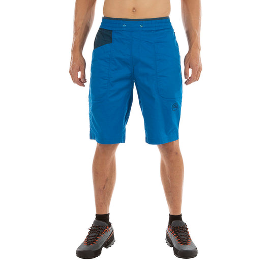 A person wears La Sportiva Bleauser Shorts Men's—bright blue, organic cotton with gusseted crotch and pockets—and black hiking shoes, standing against a white background. Only their body from neck to feet is visible.