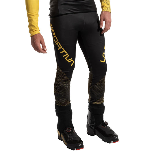 A person wears La Sportiva Stratos VI Racing Pants Men's in black and yellow with black buckled mountain boots, standing against a white background. Only the lower torso and legs are visible.