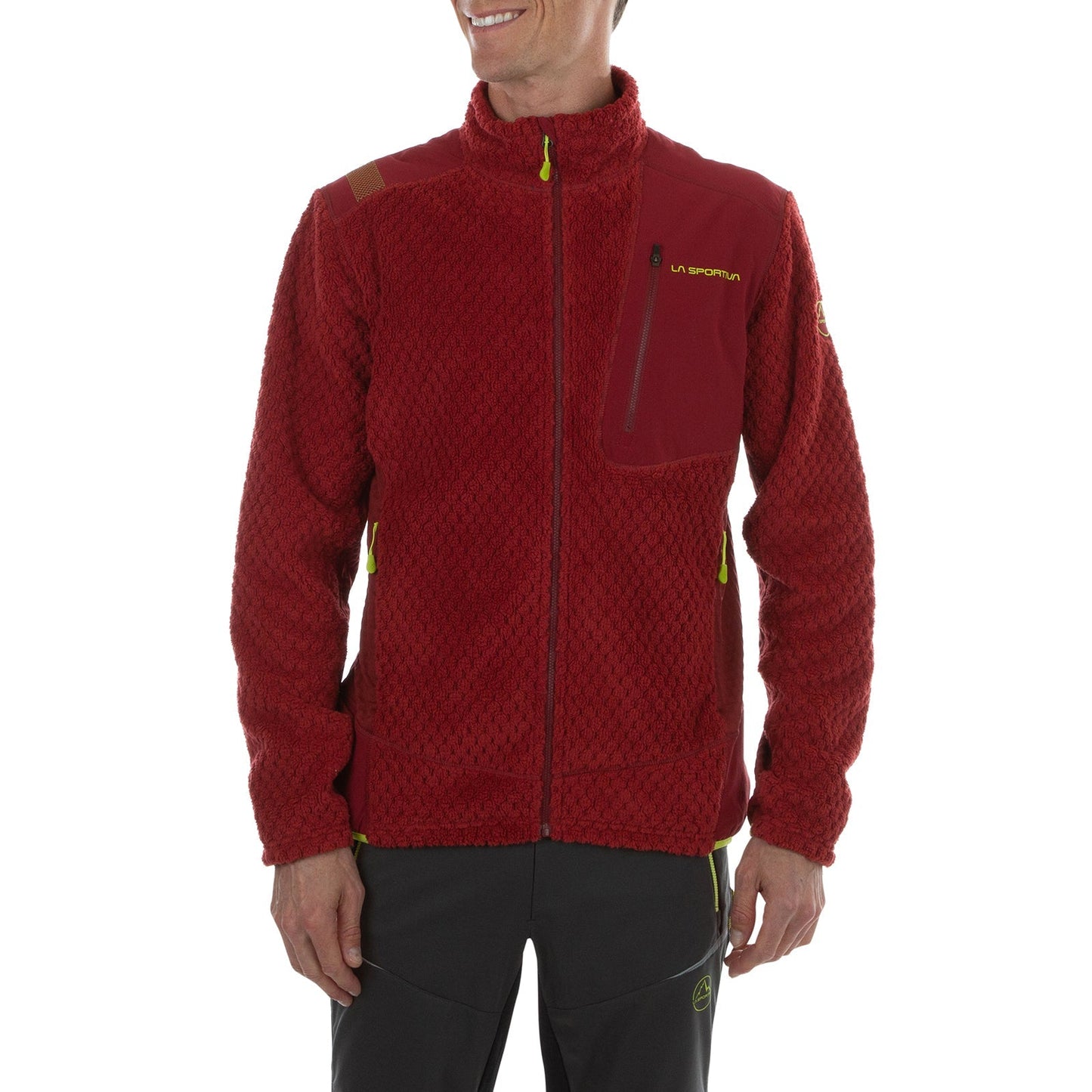 A man stands smiling with hands at his sides, wearing the La Sportiva Bristen Thermal Jacket Men's—a textured red high-loft fleece with a high collar, chest pocket, and yellow zipper accents—paired with dark pants.