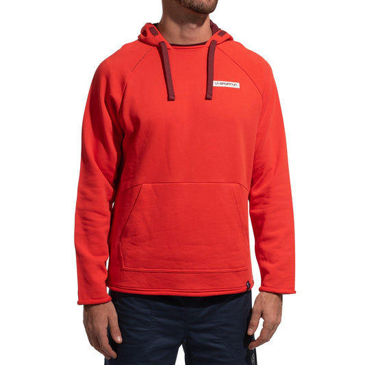 A man stands against a plain white background, wearing the La Sportiva Telendos Hoody Men's (2025) in bright red organic cotton with a front pocket, dark drawstrings, and a small rectangular logo on the left chest, paired with dark pants.