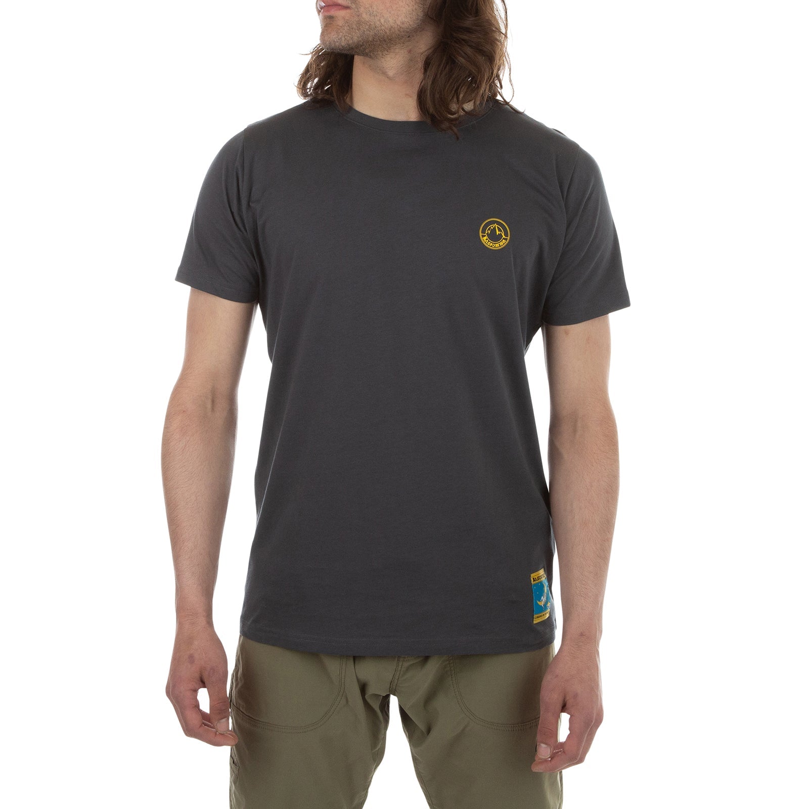 A person with long brown hair wears a La Sportiva Climbing On The Moon T-Shirt Men's (2025) in dark gray organic cotton with a small yellow logo on the left chest and khaki pants, standing against a white background for a retro vibe.