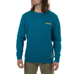 A person with long hair wears a teal La Sportiva Climbing On The Moon Sweatshirt Men's (2025) made from natural fiber, featuring a small yellow logo on the left chest and a yellow hem patch, paired with khaki pants against a white background.
