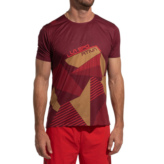 A man models the La Sportiva Comp T-Shirt Men's (2025), featuring a maroon and mustard geometric pattern with brand text, paired with red shorts, standing against a white background.