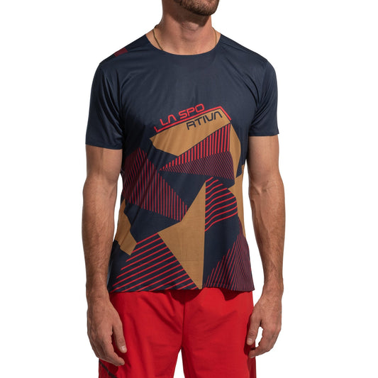 A man wears the La Sportiva Comp T-Shirt Men's (2025), a short-sleeved navy blue and tan lightweight climbing top with red accents, geometric patterns, and logo on the front, paired with red shorts against a plain white background.