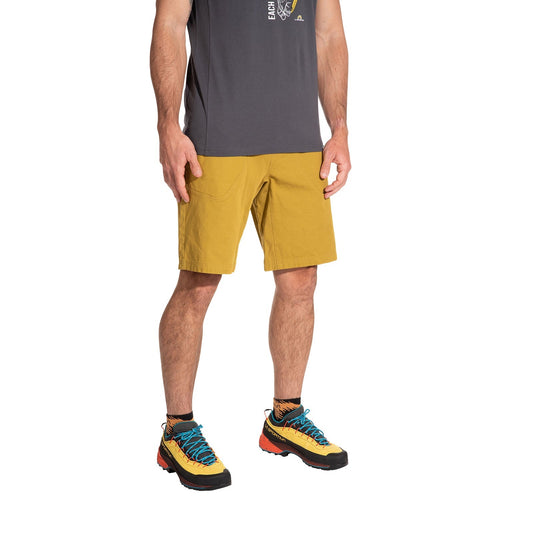 A man stands against a white background wearing La Sportiva Flatanger Shorts Men's in mustard yellow, paired with a dark gray t-shirt, black socks, and yellow hiking shoes with blue and orange accents. The outfit is designed for freedom of movement; his face is not visible.