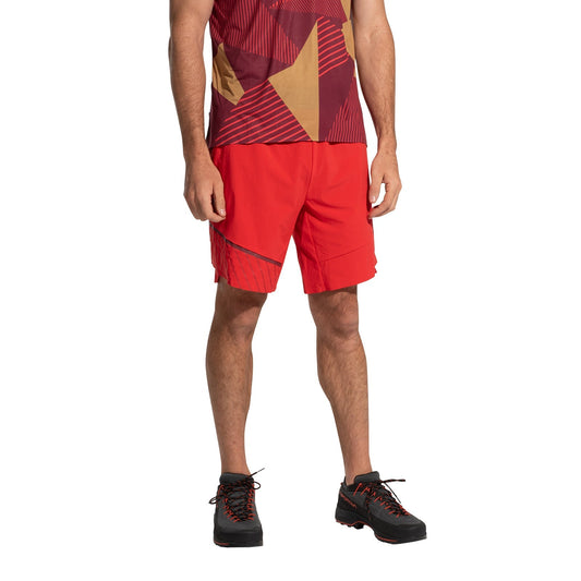 A person wears the La Sportiva Comp Shorts Men's (2025)—bright red, lightweight, and durable climbing shorts—with a red geometric-patterned sports shirt and black trail shoes with red laces, standing against a white backdrop; face not shown.