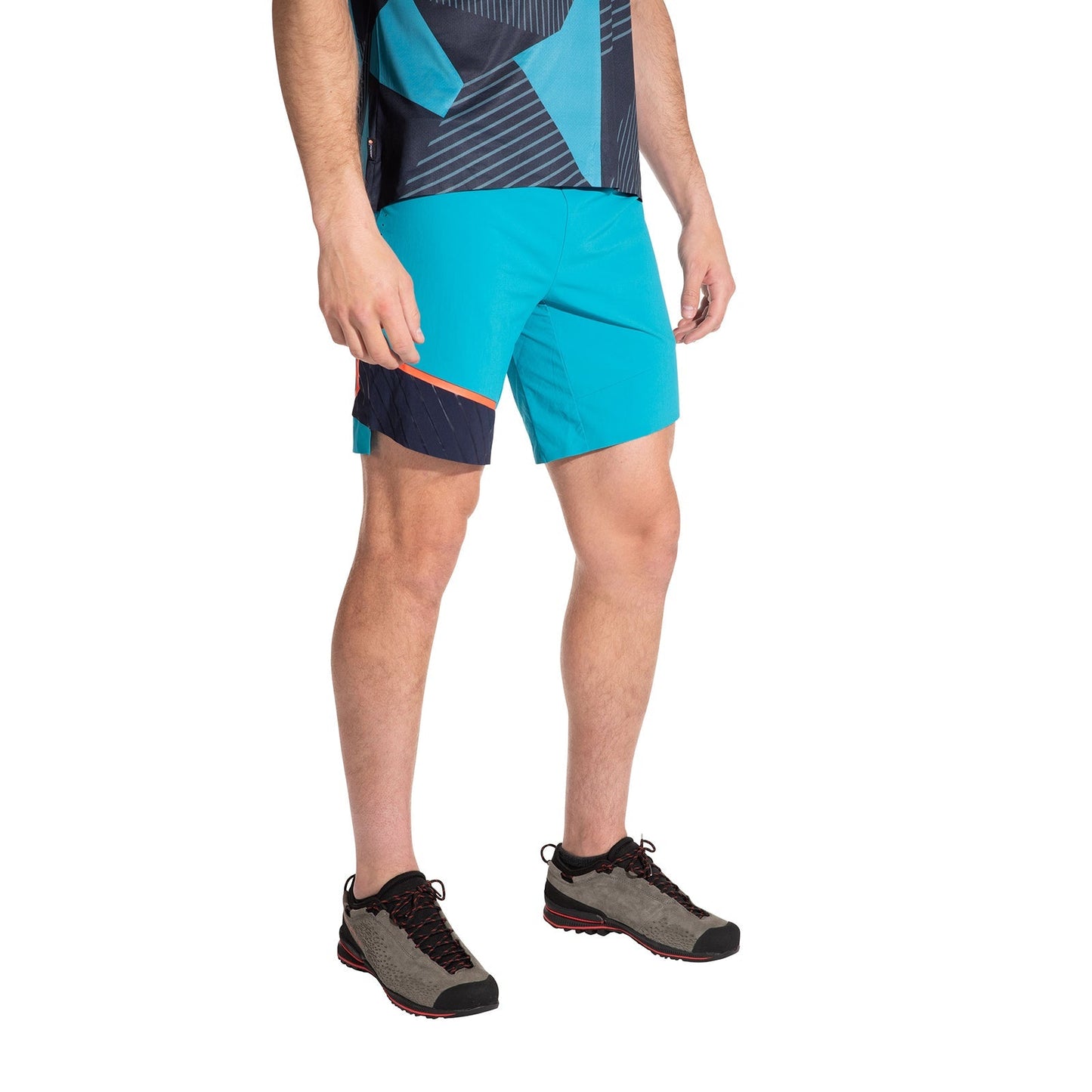 A man wearing La Sportiva Comp Shorts Men's (2025) in turquoise with navy and orange accents, paired with a geometric blue shirt and gray-black trail running shoes, stands against a white background.