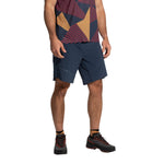 A person wearing a geometric-patterned shirt, black socks, durable black trail shoes, and La Sportiva Comp Shorts Men's (2025) in lightweight blue stands with hands relaxed at their sides; their head is not shown.