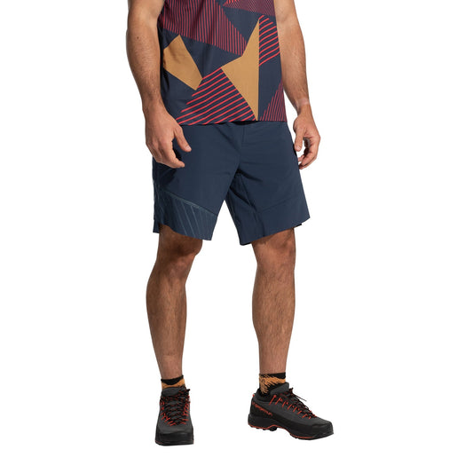 A person wearing a geometric-patterned shirt, black socks, durable black trail shoes, and La Sportiva Comp Shorts Men's (2025) in lightweight blue stands with hands relaxed at their sides; their head is not shown.