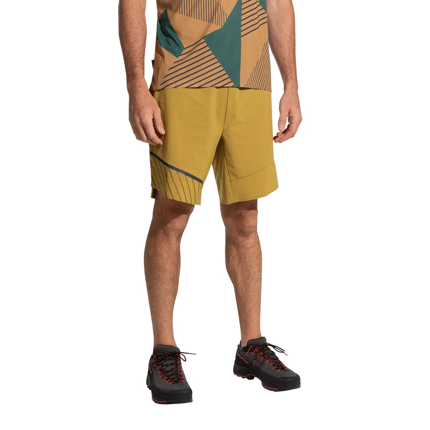 A person wears La Sportiva Comp Shorts Men's (2025) in mustard yellow with gray accents, a geometric-patterned brown and green shirt, and lightweight black hiking shoes with red laces, standing against a white background.