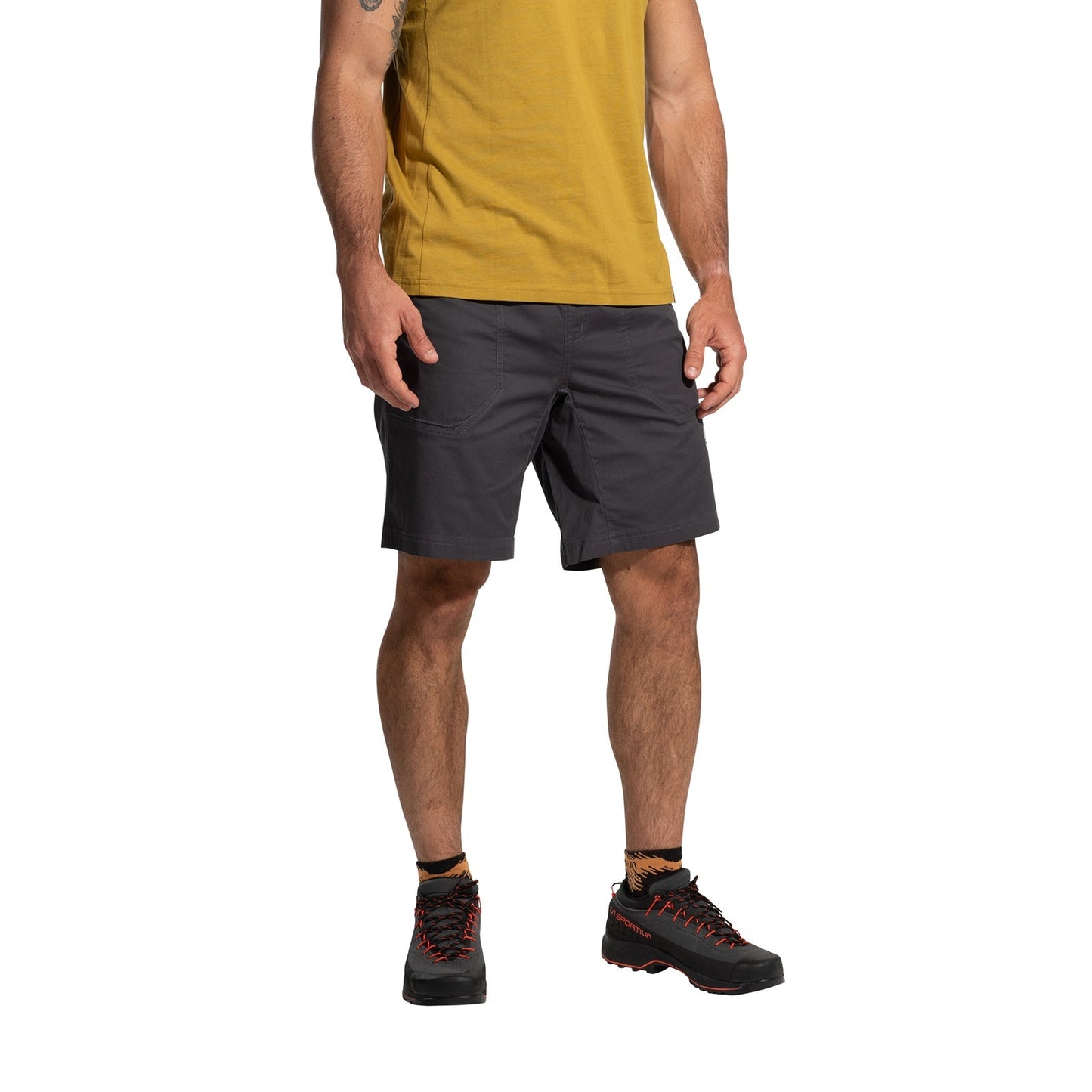 A person stands against a white background wearing a mustard yellow t-shirt, La Sportiva Roots Shorts Men's (2025) in dark gray with a comfortable waistband, and black hiking shoes with orange laces. The head is not visible in the image.