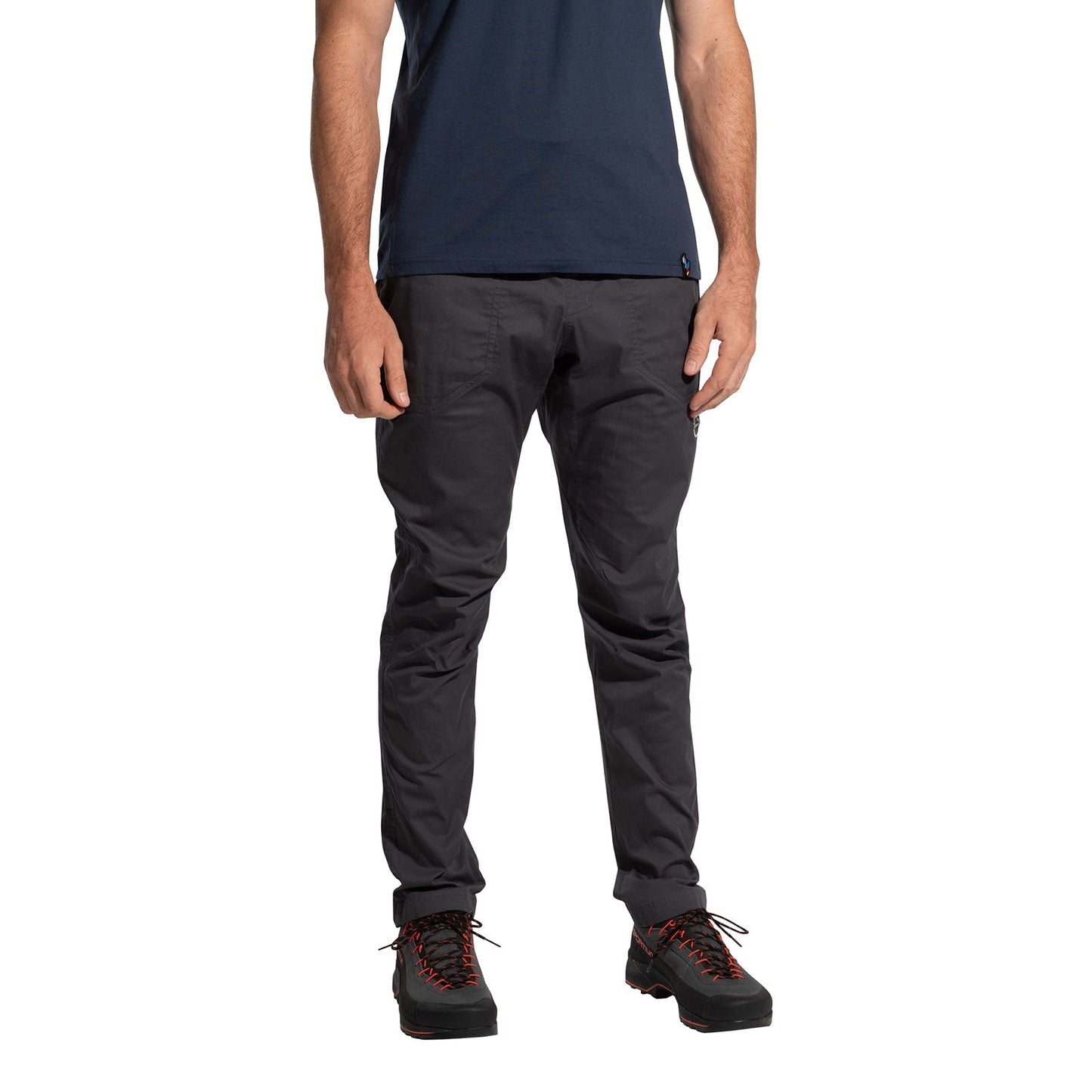 A person stands against a white background, wearing La Sportiva Roots Pants Men's (2025) in dark gray, paired with a navy t-shirt and black hiking shoes with orange laces. The minimalistic image is cropped from the shoulders down.