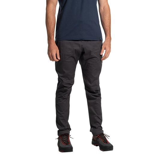 A person stands against a white background, wearing La Sportiva Roots Pants Men's (2025) in dark gray, paired with a navy t-shirt and black hiking shoes with orange laces. The minimalistic image is cropped from the shoulders down.