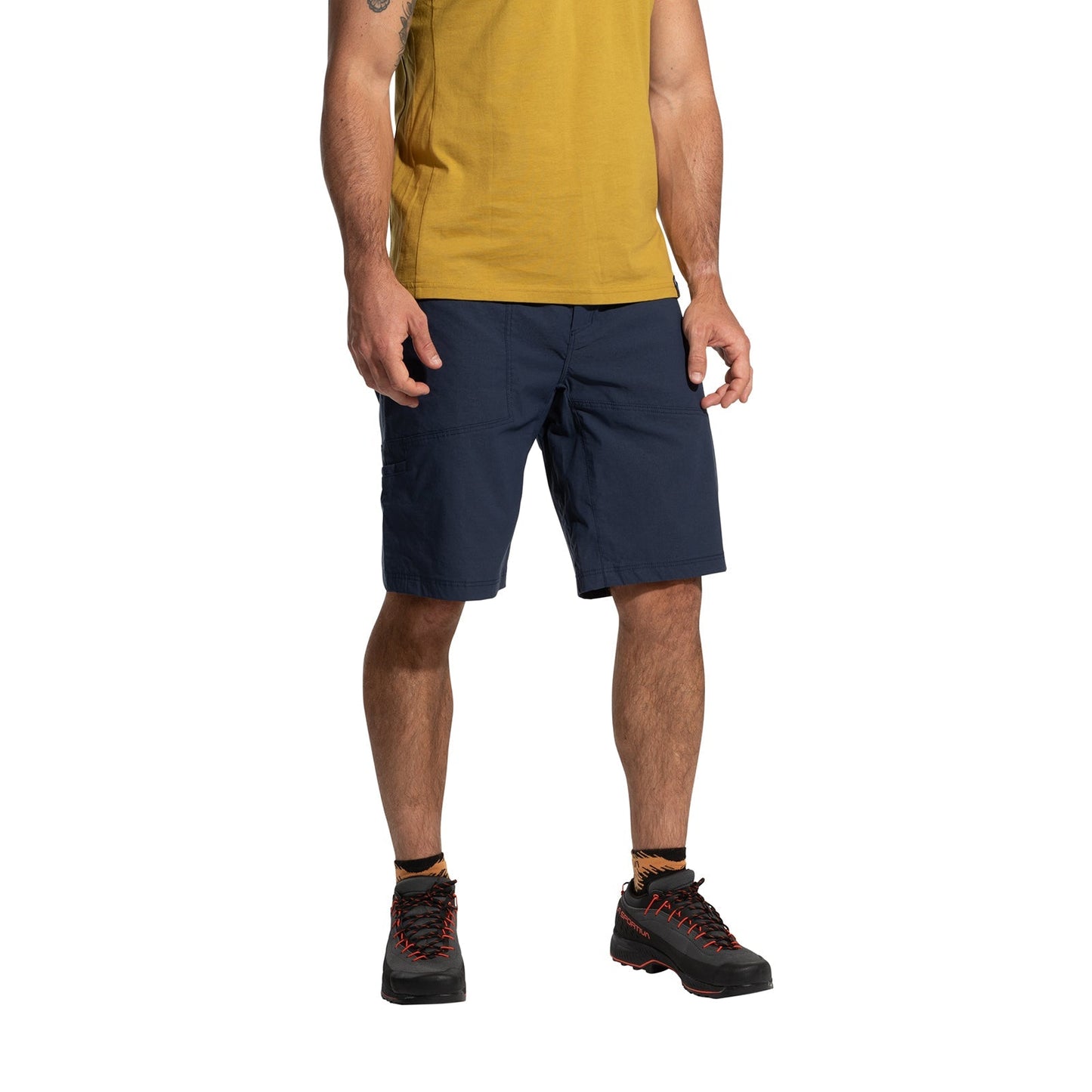 A man stands against a plain white background, wearing a yellow T-shirt, La Sportiva Talus Shorts Men's (2025) in navy blue with a chalk bag loop, and black hiking boots with orange laces. His head is not visible in the image.