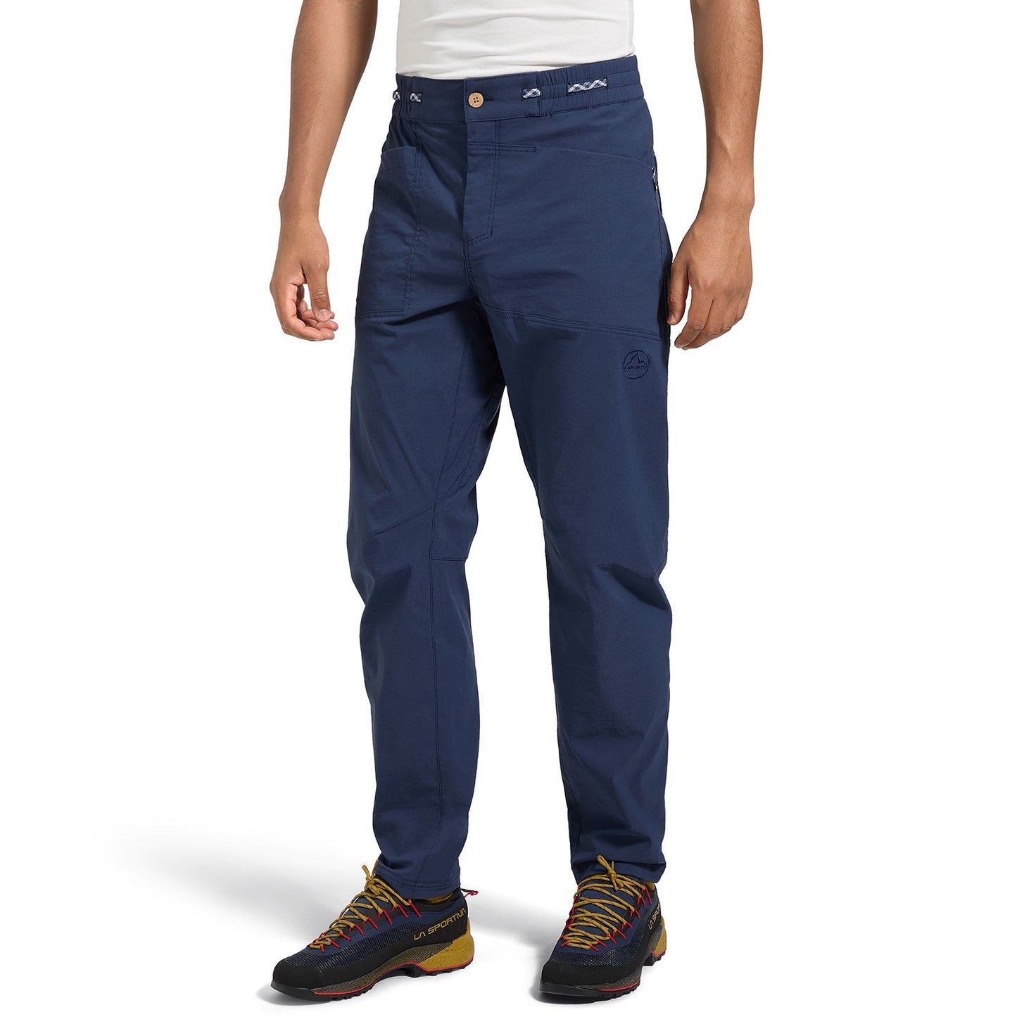 A person wearing La Sportiva Talus Pants Men's (2025) in navy blue and black hiking shoes with yellow and red accents stands against a white background, showing only the lower half of the body.